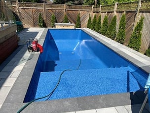 Empty rectangular swimming pool with blue water and cleaning equipment, surrounded by concrete deck and wooden privacy fence